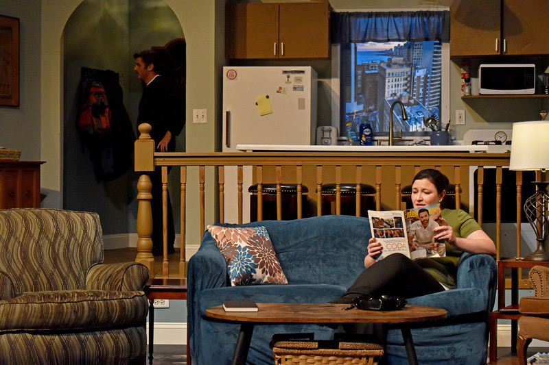 The apartment: she reads a magazine on the couch while he stands in the doorway.